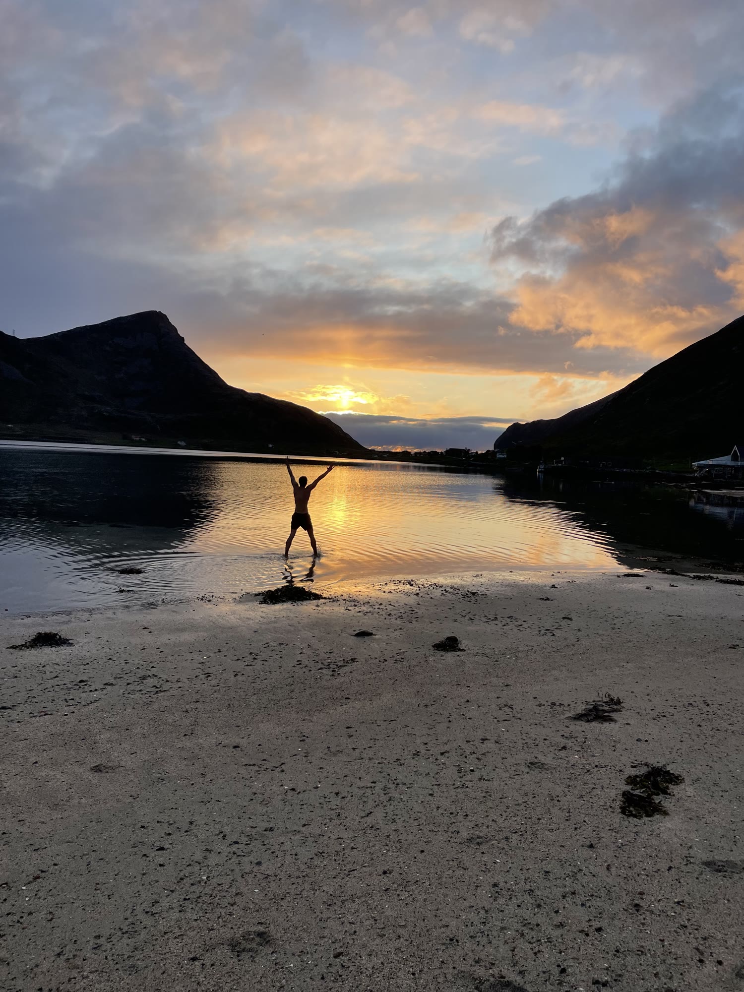 A swimmer raises both arms to the midnight sun over a still beach.