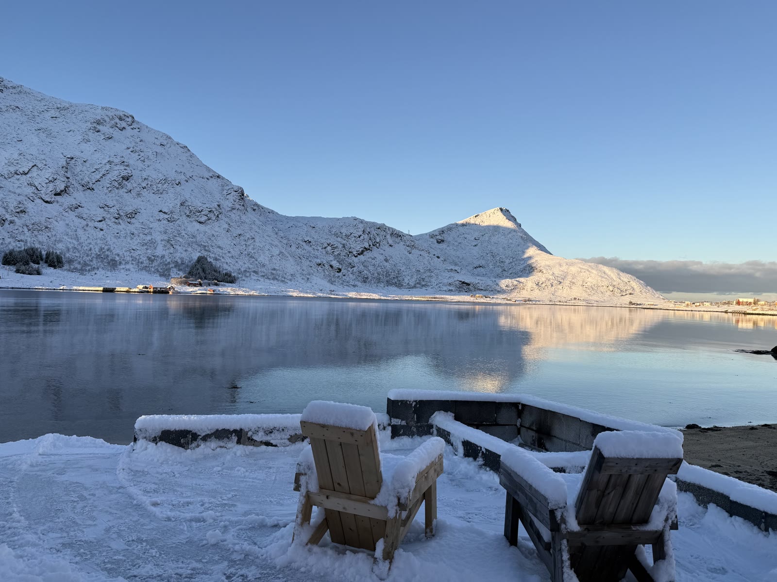 Snow-covered chairs on the dock facing a calm fjord and white winter peaks.