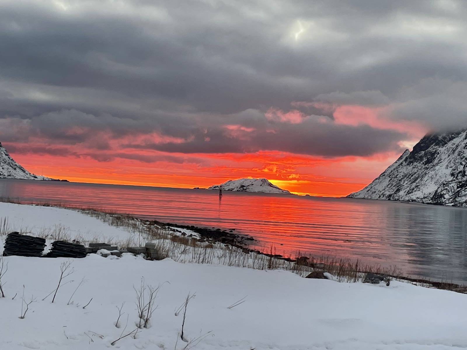 A fiery polar-night sky over a snowy fjord shore at midday in December.