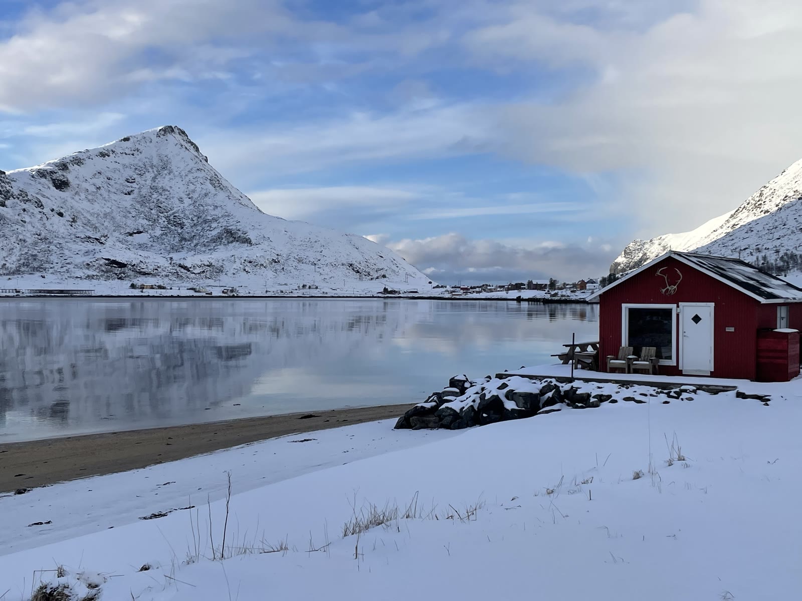 A red cabin by a calm fjord under snowy peaks and blue sky on a bright winter day.